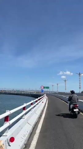 The video shows a motorcycle rider on a bridge. The rider is wearing a gray jacket and riding a white scooter. The bridge has a white railing and a concrete barrier. The water on the left side of the bridge is blue. There are traffic signs on the left side of the bridge, including a speed limit sign that says 40. The sky is blue with some clouds. There are construction cranes in the distance.