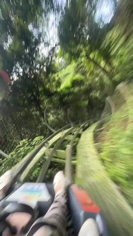 The video is a first-person perspective of a person riding a bobsled or roller coaster through a lush, green forest. The camera is mounted low, showing the front of the bobsled, the person's legs in striped pants and white shoes, and the tracks ahead. The bobsled is moving quickly, causing the surrounding trees and foliage to blur. The track has several curved sections, and the person on the ride exclaims "A turn, a turn!" in Russian. The environment appears to be a natural setting with dense vegetation and trees.