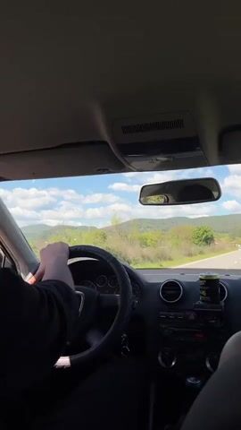 A car travels along a road. The view is from the driver's perspective. The driver is male and wears black. An energy drink sits on the dashboard. In the passenger seat is a hand wearing rings. Trees and rolling hills can be seen through the windshield. The sky is blue with clouds. There are other vehicles on the road. Electronic music plays.