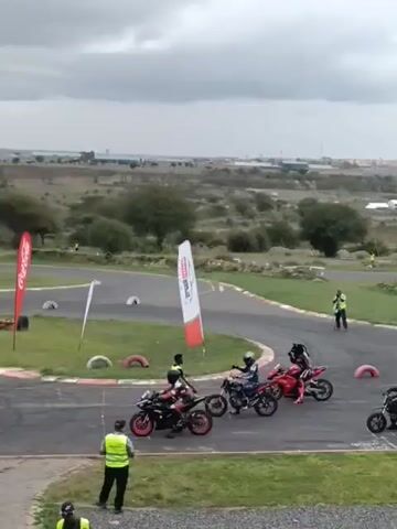 Here is a description of the video:

The video shows a group of motorcyclists at a track event. Six riders are lined up at what appears to be the starting line, each on their motorcycles. They are being attended to by race officials who wear bright yellow vests. A woman with a green flag stands off to the side, preparing to start the race. Behind the motorcycles is a grassy area with tires lining the track, a red-and-white flag, and other race officials. In the background, the terrain slopes downwards, revealing trees, bushes, and buildings in the distance under a cloudy sky.