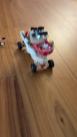 A homemade, somewhat crude, monster-like vehicle with four wheels and a menacing face is being pulled across a wooden floor by a small figure. The monster has googly eyes, sharp teeth, and a red and white body. The figure being pulled is black and white with arms and legs outstretched, and it is connected to the monster by a pink string. The monster appears to be made of cardboard and other craft materials. The scene is filmed from a low angle, emphasizing the monster's movement.