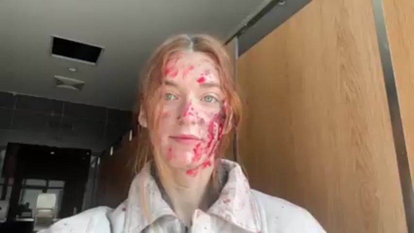 Two young women are standing indoors, apparently in a bathroom, and taking a selfie. Both women have red substances all over their faces. The woman on the left is wearing a neon yellow vest. The woman on the right is wearing a white button-down jacket. Both women are facing the camera, making slightly exaggerated expressions.