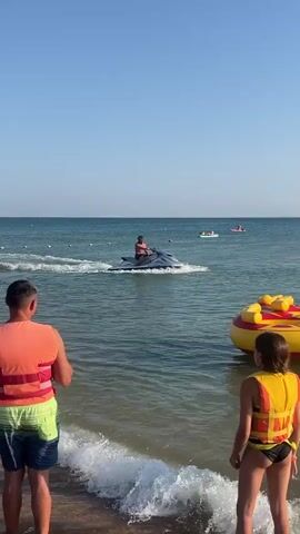 A man is riding a jet ski in the ocean. He is wearing dark shorts and a dark face covering. The jet ski is silver and gray. In the background, there are people on paddleboards and inflatable rafts. In the foreground, there are two people, one in an orange and yellow life vest and another in a yellow life vest. There is an inflatable tube with red and yellow markings in the water. The sky is clear and blue.