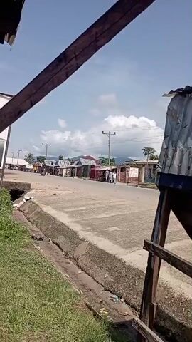 The video shows a street scene with small buildings lining one side of a road. There are a few people walking on the sidewalk and a motorcycle is passing by. The sky is blue with some clouds, and there are trees and hills in the background. The foreground shows a partial view of a wooden structure with a corrugated metal roof, possibly under construction or in disrepair.