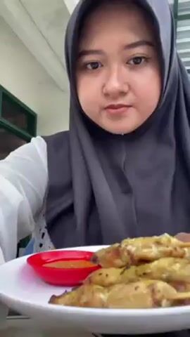 A woman wearing a white top and a gray hijab is holding a white plate of food. The plate contains several skewers of satay, a small dish of sauce, a slice of lime, and some sliced red onions. The woman takes a skewer from the plate. The video seems to be taken in an outdoor setting with a light-colored roof and a green window frame visible in the background. The woman is the main focus, and the video primarily showcases the food she is about to eat.