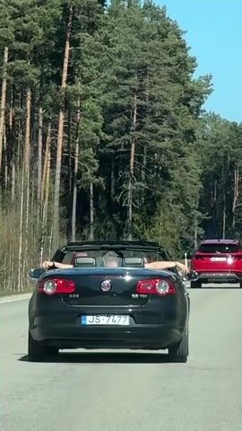 A black convertible car drives down a road lined with tall trees, followed by a red car. The passenger of the convertible has their arms raised and resting on the back of the driver's seat. The convertible has a license plate that reads "JS-7477". The sky is blue and clear.