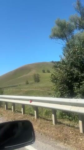 The video shows a view from a vehicle moving along a road. In the foreground, a car's side mirror is visible. A white guardrail runs alongside the road. Beyond the guardrail, a grassy hillside slopes upwards towards a clear blue sky. Scattered trees dot the hillside, and a small building with a white roof and fence can be seen partway up the slope. The landscape appears to be rural and serene under bright daylight.