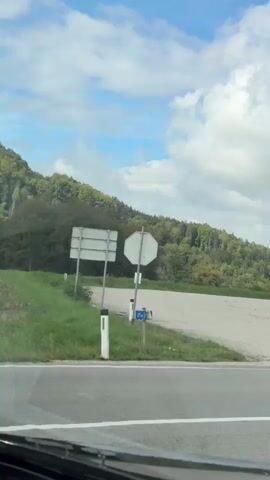The video opens with a view of a paved road with white lane markings. In the background, there is a sloping hill covered in green trees under a blue sky with white clouds. The camera then pans to the right, showing a white building with signage. A white car is seen turning, leaving tire marks on the asphalt. The video continues to show a stretch of road with green grass on the side, bordered by a line of trees. Further down the road, other cars are visible. The building on the right has a prominent red section and a long row of windows. A traffic mirror is mounted on a pole in front of the building. The scene appears to be a semi-rural or suburban area with some commercial buildings and natural landscapes. The weather is overcast with patches of blue sky.