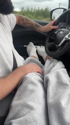 The video shows the interior of a car from the passenger's perspective. A person with tattoos on their left arm is driving. Their right hand is on the steering wheel. The driver's lap is visible, and the passenger's legs are extended in front of them, wearing grey sweatpants and white socks. The passenger's feet are visible, one resting on the dashboard and the other near the pedals. The dashboard displays a stereo system, a hanging air freshener, and a small religious icon. The view outside the car window shows a road with other vehicles and a cloudy sky. The video appears to be taken while driving.