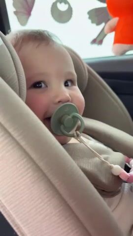 A baby is seen from the chest up, seated in a light-colored car seat, likely in the back of a car in Staroderevyankovskaya, Russia. The baby has light hair and dark eyes and is wearing a light-colored outfit, possibly ribbed. A green pacifier is in the baby's mouth, attached by a string with white and pink beads.

In the background, hanging decorations are visible: a white butterfly, a white bib-shaped item, and an orange fox plush toy. The lighting suggests it is daytime.

Throughout the short video, the baby looks directly at the camera, displaying a range of expressions. At the beginning (0:00), the baby is smiling faintly. This expression shifts to a more serious or contemplative look around 0:01, before returning to a subtle smile towards the end (0:04-0:05). The baby's head can be seen moving slightly, appearing to bob to the rhythm of the Russian music playing in the background. No visible text is present in the video.