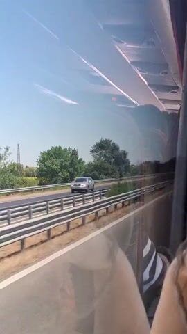 The video captures a view from a moving vehicle, likely a bus, traveling on a highway. The perspective is from a side window, showing the passing scenery. On the left side of the frame, a highway stretches ahead, bordered by a metal guardrail. Beyond the guardrail, there are fields of yellow flowers, likely sunflowers, interspersed with trees and greenery. A tall industrial structure, possibly a power line tower, is visible in the distance. Several cars are seen on the highway, including a silver SUV and a gray Mini Cooper Clubman in the foreground. The sky is clear and blue, suggesting a sunny day. Reflections from inside the vehicle are visible on the window, obscuring parts of the view and showing interior elements like a seat and possibly a digital display showing the temperature as 28 degrees Celsius. The overall impression is a travel scene on a warm, bright day.