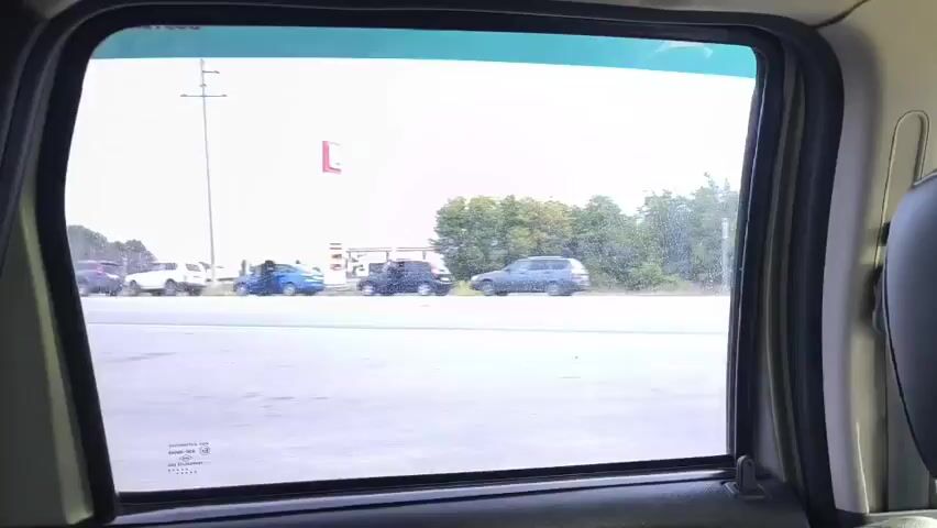 The video shows a view from inside a vehicle, looking out a window at a road with cars parked on the side. There are trees and vegetation behind the parked cars. In the first frame, there's a gas station in the distance. In subsequent frames, people are seen near some of the parked cars. The weather appears overcast. The video captures a sequence of parked vehicles and roadside scenery as the camera's perspective moves forward.