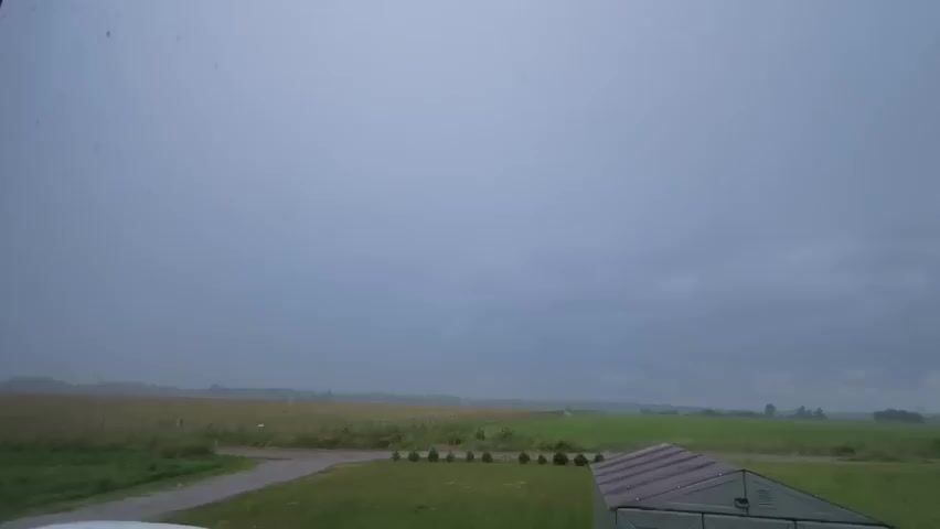 The video shows a wide shot of a cloudy, overcast sky with a hint of a storm approaching from the distance. The landscape below consists of green grass in the foreground, a gravel path, a field of yellowing crops, and more distant green fields. A dark green greenhouse with a brown roof is visible on the right side of the frame, partially cut off. The sky is a uniform gray, with no visible sun or distinct clouds, suggesting a pre-storm atmosphere or general overcast weather. There is no movement or action captured in the frame, apart from the subtle nuances of the atmospheric conditions.
