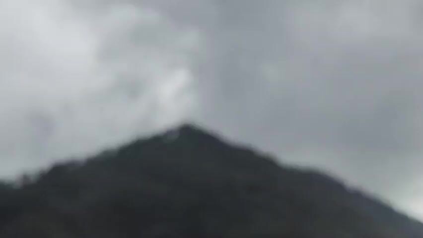 The video is a static shot of a mountain peak. The sky is overcast and appears to be filled with dark clouds. The mountain itself is covered in dark green vegetation and a line of trees runs along the very top of the peak. The image is dark and gloomy, suggesting it may be raining or about to rain. There are no people or animals visible in the video.