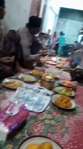 A low-angle, handheld video shot from the floor shows a gathering of people in what appears to be a home. Several plates of food are spread across a patterned rug. In the foreground, there are glasses of liquid and a box of tissues. In the mid-ground, a golden teapot sits on a tray. Several people, including men, women, and children, are seated or standing around the food. One man in the left mid-ground is wearing a black cap and a light-colored shirt and appears to be eating. In the background, there are doorways and what looks like a motorbike. The lighting is dim and appears to be coming from overhead sources.