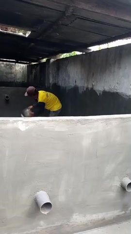 A person wearing a yellow shirt and a brown cap is applying cement to the wall of a concrete structure. The person is holding a bucket and a trowel. There are pipes protruding from the walls. The structure is covered by a corrugated metal roof. The scene appears to be an outdoor construction site.