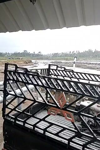 The video captures a murky, rain-soaked landscape with a river in the background and a black vehicle in the foreground. The vehicle appears to be a pickup truck with a rack on top, fitted with what looks like corrugated metal sheeting and black metal framework. The ground is muddy and uneven, with puddles of water. In the distance, a few scattered trees and some machinery, possibly excavators, can be seen. The sky is overcast, suggesting ongoing or recent rainfall. The overall impression is of a rural or semi-industrial area experiencing inclement weather.