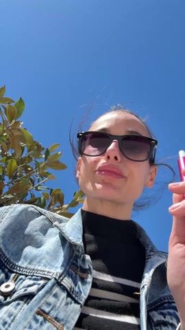 A woman is standing outside on a sunny day. The camera angle is low, looking up at her. She is wearing sunglasses, a denim jacket, and a black shirt with white stripes. She holds a purple vape pen and takes a drag from it. A tree with green leaves is visible in the background, and the sky is a clear blue.