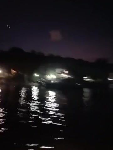 The video shows a night-time view of a body of water reflecting lights from buildings and other sources on the opposite shore. The sky is dark with a hint of purple. The water has small ripples and reflects the lights, creating bright streaks. The far shore is lined with trees and buildings.  The overall lighting is dim, and the quality of the video is somewhat shaky.