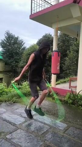 A young person wearing a black hooded vest, grey shorts, and black athletic shoes is jumping rope outdoors. The person is in motion, mid-jump, with the green jump rope blurred around them. A red punching bag hangs to the right of the frame, attached to a white pillar. The ground is paved with large, irregular stone tiles. Lush green foliage and trees are visible in the background, along with a building with a pink and white facade and a balcony railing. The sky appears overcast.