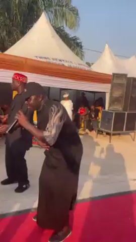 Two men are standing on a red carpet outside in front of a white tent, both holding microphones and seemingly singing or speaking. The man on the left is wearing a black tunic and pants with a red hat that says "CHUCK". He has a necklace with large beads. The man on the right is wearing a black outfit with a cap and sneakers. He is dancing slightly as he sings. Behind them, under the tent, people are seated. To the right of the men are large black speakers stacked on top of each other. The background includes palm trees and a clear blue sky.  The tent appears to have text on it that says "THINK RENTALS & EVENT".