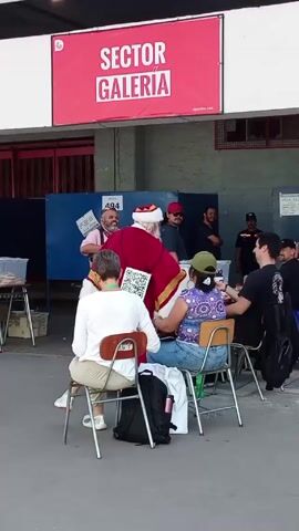 The video shows a man dressed as Santa Claus walking among people seated at desks in an outdoor setting. A red sign reading "SECTOR GALERIA" hangs above the scene. The Santa character is wearing a red and white suit. A person in a white shirt and khaki pants sits at a desk to Santa’s left. Another person in a purple patterned shirt and jeans sits at the next desk. There are several other people standing or sitting in the background. A child runs across the frame towards the camera at one point. The environment appears to be a shaded public space, possibly an outdoor market or fair.