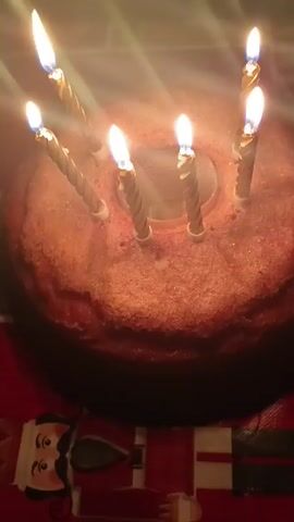 The video shows a round cake with seven lit candles on top. The cake is placed on a surface with a figure of a man in a red coat. The lighting in the room is dim, except for the light emanating from the burning candles. Someone is speaking, and there are other indistinguishable sounds in the background.