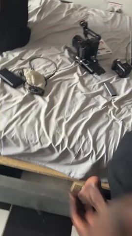 The video shows an interior shot of a room that appears to function as a recording studio and bedroom. The primary focus is a bed with disheveled grey sheets, upon which various pieces of equipment are scattered including a camera rig, microphone, external drive, laptop, and other electronics. The laptop screen is illuminated, showing what seems to be editing software. A man wearing a black vest and yellow shorts sits on the bed, facing away from the camera, using his phone. Another man wearing a "HARDEN 13" basketball jersey stands at a desk, also focused on a laptop. The room has a black-and-white tiled floor, and a fan is visible. The walls appear worn and stained. The overall impression is a cluttered workspace combined with living quarters.