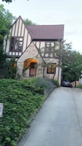 A black pickup truck is parked on a paved driveway that slopes upwards towards two houses. The house on the left is a light-colored two-story home with a red brick lower level, black shutters, and a wreath on the front door. The house on the right is a Tudor-style home with a light-colored stucco exterior, dark trim, stone accents, and ivy growing on the side. A tree is situated between the two houses. The grass in front of the houses is neatly manicured, and some low landscape lighting is visible on the lawn. The sky is overcast, and the time of day appears to be late afternoon or early evening.