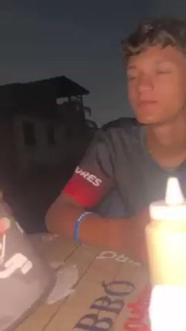 A video depicts two young men seated at a table outdoors. The table is covered with a printed design and holds various condiments in squeeze bottles, including white, yellow, and red. One of the men is wearing a dark-colored t-shirt with a red sleeve, while the other is wearing a white t-shirt. Initially, both men appear relaxed, but the second man uses a spatula to flip a portion of food toward the camera before putting his face up to the camera. The background is dark, hinting at nighttime. Buildings and possibly trees are visible in the distance.