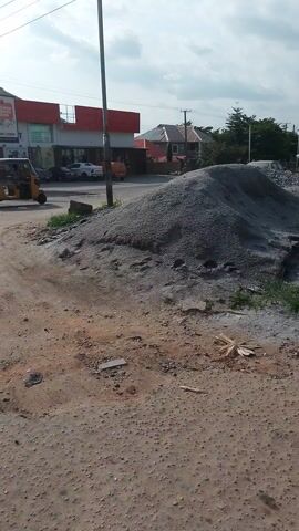 The media file depicts a street scene in Dutsen Alhaji, Nigeria, likely during daytime under cloudy skies. The perspective is from a moving vehicle, showing a road under construction or repair. A large pile of gravel is visible on the right side of the frame, indicating ongoing work.

In the foreground, a section of a road surface made of loose gravel and dirt is prominent. To the left, a tall metal pole with electrical wires stretches upwards, a common sight in many urban and semi-urban areas. Further down the road, several vehicles are visible: a yellow auto-rickshaw or Keke Napep, a dark car, and a red car. These vehicles suggest typical street traffic.

Adjacent to the road, on the left, are buildings. One appears to be a commercial establishment with a red and white facade and prominent signage, though the text is not clearly legible. Next to it, another building is under construction, characterized by extensive scaffolding made of bamboo poles. This construction site also features exposed brickwork and concrete elements, suggesting a multi-story structure is being built. A large black water tank is also visible near the construction site.

The overall scene suggests a developing urban or semi-urban environment with ongoing infrastructure work and commercial activity. The presence of construction and the type of vehicles are indicative of Nigeria. The weather appears overcast, which could mean it's morning or afternoon with cloud cover. There are no visible people prominently featured in the frame, but their presence is implied by the vehicles and the context of a street. The activity is primarily centered around road maintenance and construction, with traffic passing by.