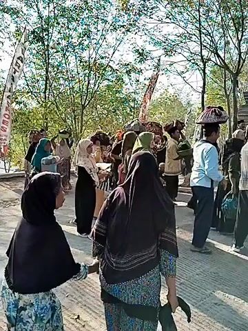 A group of people, mostly women wearing headscarves and traditional clothing, are walking in procession outdoors. Many are carrying items on their heads, such as baskets or fabric. They are walking on a paved surface, possibly a road or pathway, with trees and foliage in the background. There are also flags or banners visible. The weather appears to be sunny. The overall scene suggests a cultural event or ceremony.