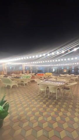 The video shows an empty outdoor seating area of a restaurant at night. The area is paved with a geometric pattern of red, yellow, and brown tiles. String lights are hung overhead, illuminating the space. White wicker tables and chairs are arranged throughout the area. In the background, there are more tables and chairs under a covered area, and some greenery is visible. The overall atmosphere is calm and quiet.