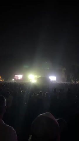 This is a low-light video of a large outdoor gathering at night, possibly a concert or festival. A brightly lit stage with screens is visible in the distance, with spotlights cutting through smoke or mist. A dense crowd of people are gathered in front of the stage, their faces mostly obscured by darkness. The overall atmosphere appears to be one of excitement and festivity, with a sense of shared experience among the attendees.