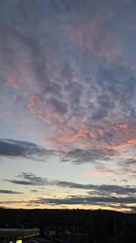 The video is a time-lapse of a sunset over a landscape of trees and buildings. The sky is filled with clouds that are tinged with pink and orange hues. The sun is setting on the horizon, casting a warm glow over the scene. The video pans slightly upwards to capture the full expanse of the sky. There are no people or specific actions depicted in the video, it is purely a scenic time-lapse.