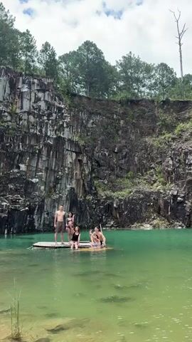 A group of five people are gathered on a makeshift raft in a quarry. The raft is made of logs lashed together and is floating on clear turquoise water. The quarry walls are steep and rocky, with trees growing at the top. The people are dressed in swimsuits. One man is standing on the raft and raising his arm in the air. Another man is sitting on the raft and looking at the camera. Three women are sitting on the raft. The water is calm and reflects the sky. The scene appears to be a recreational outing.