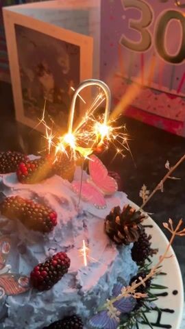 A close-up shot features a birthday cake decorated with light blue frosting, blackberries, pine cones, and butterfly ornaments. A sparkler in the shape of the number "0" emits bright sparks atop the cake. In the background, a "30" birthday card stands along with another card featuring a framed photograph.  The cake sits on a white plate with a black design along the edge. The sparkler is actively burning.