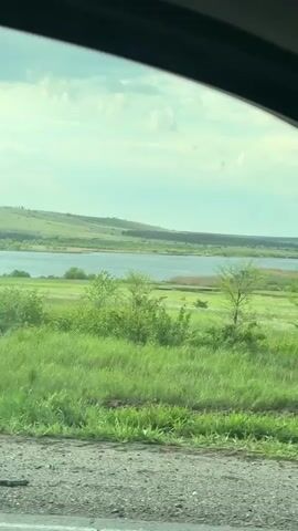 The video shows a view from a car window of a lake surrounded by green fields and hills. The sky is cloudy and bright. The road is visible in the foreground. The car appears to be moving.
