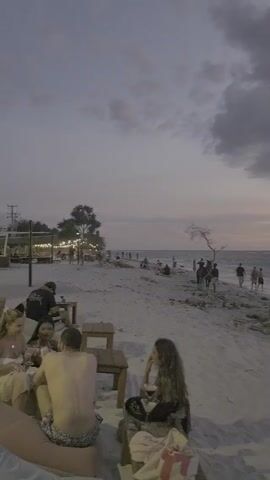 The video shows a sandy beach scene at dusk with people scattered around.  In the foreground, a shirtless man and a woman sit at beach furniture.  The woman holds a glass and looks around. In the midground, other people walk or stand near the water's edge. The water appears choppy with small waves.  Several trees, some leaning towards the ocean, stand along the shoreline. In the background, more people gather near a structure lit with string lights. The sky is overcast with gray clouds and a hint of sunset color near the horizon. The overall tone is somewhat muted.