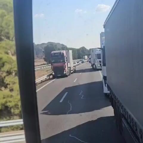 A first-person perspective video shows a highway scene with several trucks and a black car. The camera is mounted inside a vehicle, likely another truck given the close proximity to the adjacent vehicle's trailer. In the foreground, the side of a large white truck trailer is visible on the right. The highway stretches ahead with two lanes visible. On the left lane, a red and black truck is prominently displayed, followed by a black car. Further down the highway, additional trucks are visible, suggesting a traffic jam or slow-moving traffic. The environment appears to be rural with dry vegetation and hills in the background under a clear blue sky. The sun is casting shadows, indicating it's daytime. The video seems to be a recording of a traffic situation on a highway.