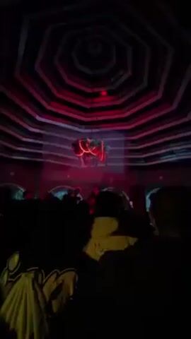 The video depicts a performance by a male singer on a small stage, seen from within a crowd of attendees. The venue features a distinctive ceiling design with concentric black and white striped patterns, illuminated with red lighting. A decorative chandelier-like structure hangs above the singer. The singer, who is wearing sunglasses, is rapping into a microphone and gesturing with his hand in the air, while audience members record the performance on their phones. The lighting in the venue is dim.