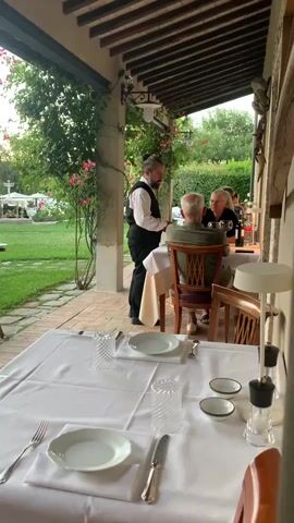 A waiter in a white shirt and black vest stands to the left of a table, facing two people sitting at a dining table on an outdoor patio. The table is set with white tablecloths, plates, silverware, and glasses. To the left of the table, a lush green lawn is visible, with rose bushes and trees. In the background, more tables and umbrellas are visible, suggesting a restaurant or outdoor dining area. The waiter appears to be interacting with the diners.