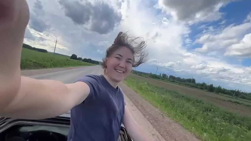 A young woman with her hair pulled up in a bun smiles as she leans out of a car window. She is wearing a dark blue t-shirt. The car is moving, as indicated by the blurred background and wind blowing her hair. The scene takes place on a road bordered by green fields and trees under a cloudy blue sky.