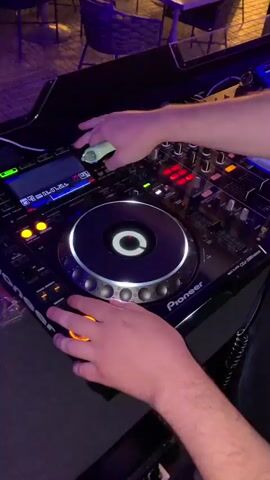 A close-up, high-angle shot shows a person's hands operating a DJ setup. The DJ is wearing a light-colored shirt. The left hand is on the jog wheel of a Pioneer CDJ-2000 Nexus 2, while the right hand holds a white object and manipulates a knob on the mixer. The CDJ's screen displays track information. The mixer has red and white buttons and sliders. The background is blurred, showing what appears to be a dimly lit venue with tables and chairs, possibly an outdoor patio area with string lights. The lighting is a mix of blue and purple hues, creating a club-like atmosphere. The overall scene depicts a DJ in the process of mixing music.
