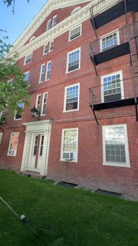 The video pans upward and to the right, showing the brick exterior of a multi-story building, likely a dormitory or apartment building. The building has numerous windows with white frames and some have air conditioning units installed. There are also several black metal fire escapes attached to the side of the building. The building is surrounded by green grass and a tree with green leaves is visible on the left side of the frame. The sky is blue and clear. There are no people visible in the video. The overall impression is of an older, well-maintained building in an academic or residential setting.