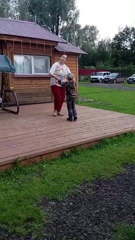 A woman in a white shirt and red pants and a child in a camouflage outfit are dancing on a wooden deck in front of a wooden house. The woman is holding the child's hands and they are moving in a circular motion. The child is facing away from the camera. The deck is surrounded by green grass. In the background, there are trees, a red fence, and several parked cars. The weather appears to be overcast.
