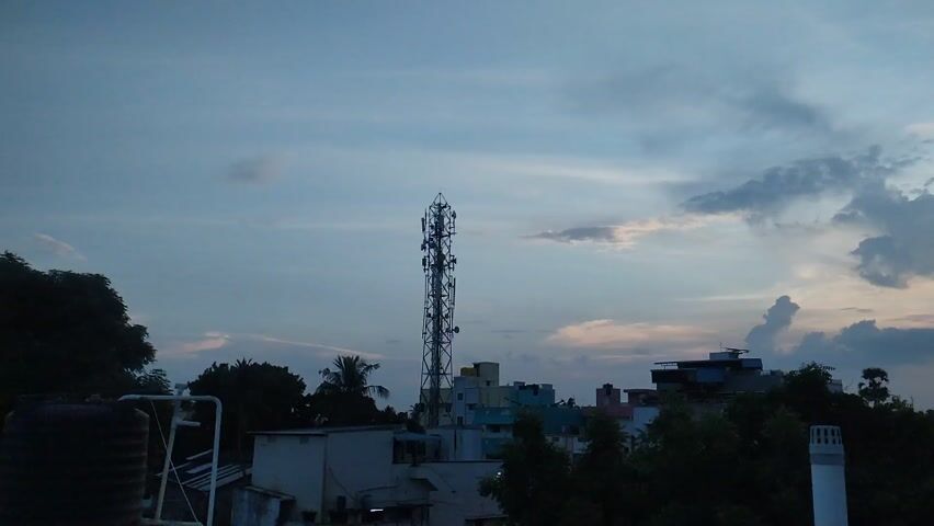 The media file captures an evening scene in Maduravoyal, India, as dusk settles. The sky is a canvas of soft blues and grays, with scattered clouds tinged with the warm hues of a setting sun, suggesting a pleasant weather condition. Dominating the landscape are two tall, lattice-like telecommunication towers, one prominently positioned on the left and another on the right of the frame. These towers, equipped with multiple antennae, are characteristic of urban and semi-urban environments where communication infrastructure is dense.

Below the towers, a cluster of buildings of varying heights and colors form the cityscape. These structures appear to be residential or mixed-use buildings, with some showing signs of being older constructions. Lush green trees and foliage create a natural contrast to the man-made structures, with dense trees filling the foreground and parts of the middle ground, hinting at a location with both urban development and significant greenery.

The overall mood is serene and tranquil, amplified by the soft, fading light of the day. The absence of people suggests a quiet moment, perhaps a typical evening in this part of India. The composition, with the towers reaching towards the sky and the buildings nestled below, creates a sense of depth. There is no visible text in the media file.