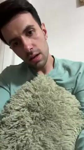 Here's a detailed description of the video:

The video shows a man pouring a bluish-green gel into his open palm. He's wearing a light green t-shirt and is sitting in front of a fluffy, light-green pillow. His face is visible, and he looks directly at the camera. He pours more gel into his hand. The cap of the tube is green. The setting appears to be indoors, possibly a living room or bedroom, with a painting visible on the wall in the background.