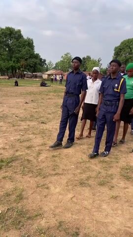 A small group of young people walk in formation across a dirt field. Two boys in dark blue uniforms lead the group. Other people, wearing various colors of clothing, follow behind them. Some of them are holding flags. The sky is partly cloudy. Trees and buildings appear in the background.