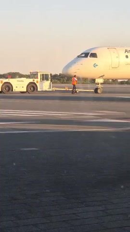 The video shows an airplane on an airport tarmac. A tow tractor is attached to the front of the airplane. A person in an orange vest stands between the tow tractor and the airplane. The airplane is white with blue and green markings. The sky is clear and the sun appears to be low in the sky, casting a warm light.